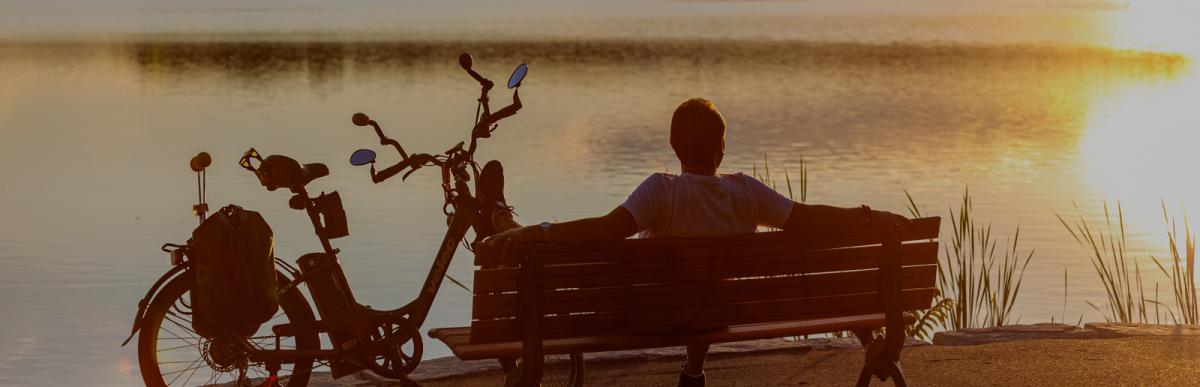 Silhouette of a man sitting on a park bench with bike looking out over water and setting sun.