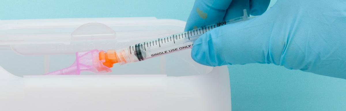 A surgically gloved hand disposing of a syringe and needle(with safety cover) being disposed of in a safe, protective container known as a Sharp's container.